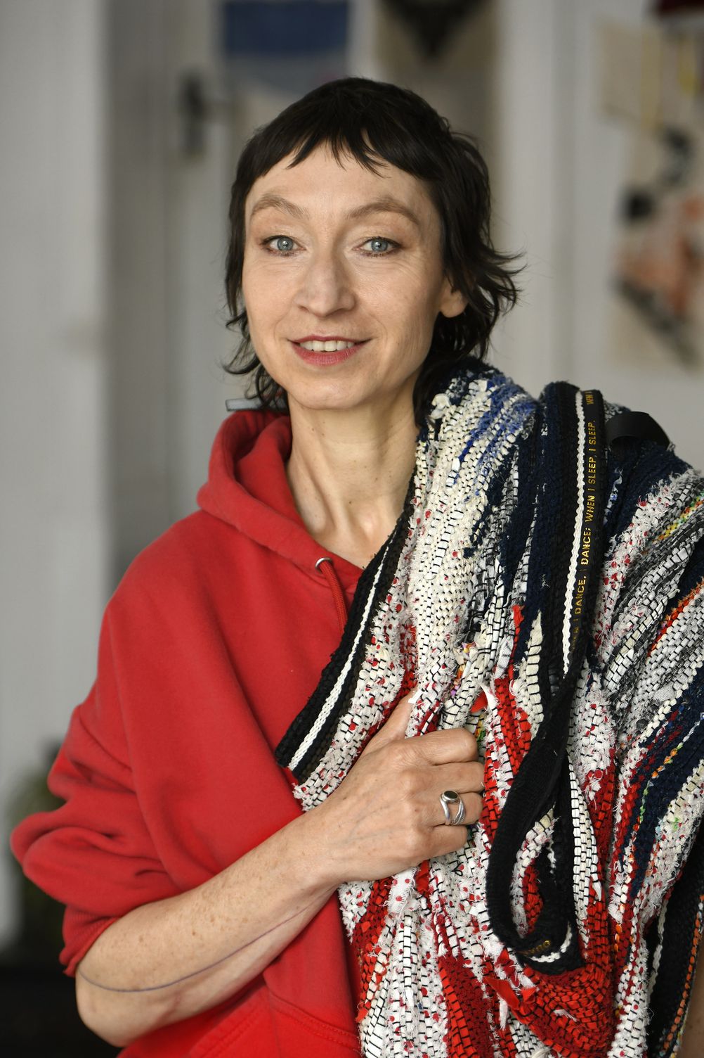 Portrait of Claudia Hill. She smiles into the camera, wearing a bright red hoodie with her poncho from Social Fabric Earth Return draped on her left shoulder.