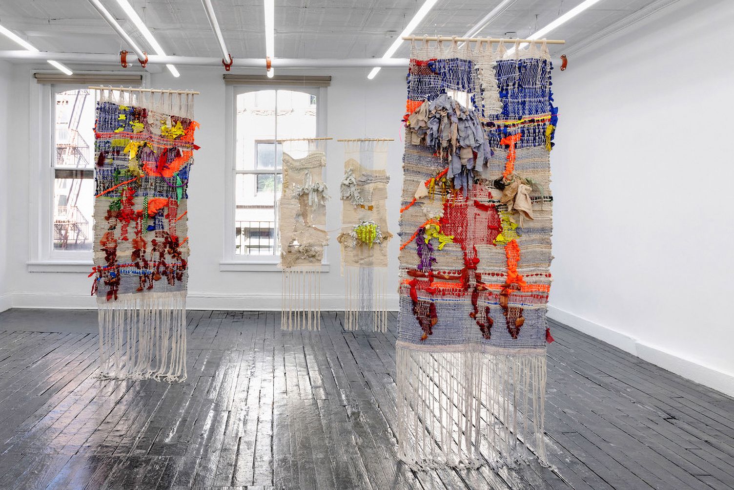 Installation view of Here We Leave Solid Ground at Kate Werble Gallery, New York: the two colourful panels of Love is as Love Does and, between them, the pale panels of Through the Clouds, hanging free above a glossy dark wooden floor