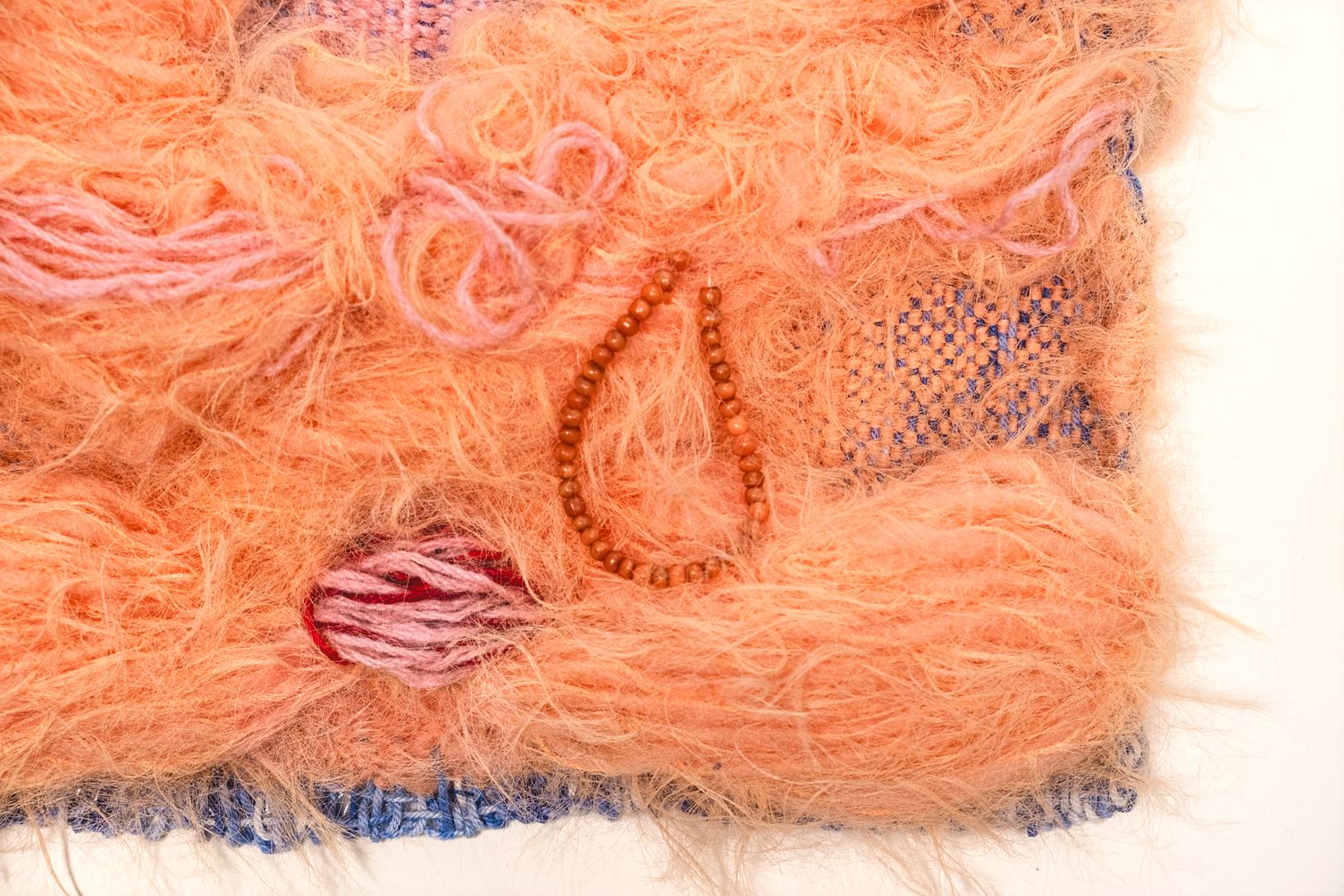 Detail of Abundance: fluffy apricot mohair with pink loops, a teardrop of wooden beads and a pink and red knot, over pink and blue weaving