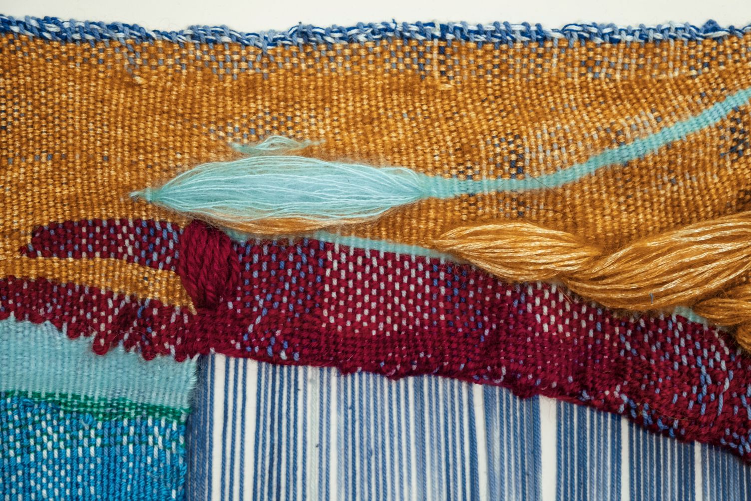 Detail of All For Us to Enjoy: a turquoise tuft and a golden twisted strand on ochre weaving, above a wine red band and open blue warp