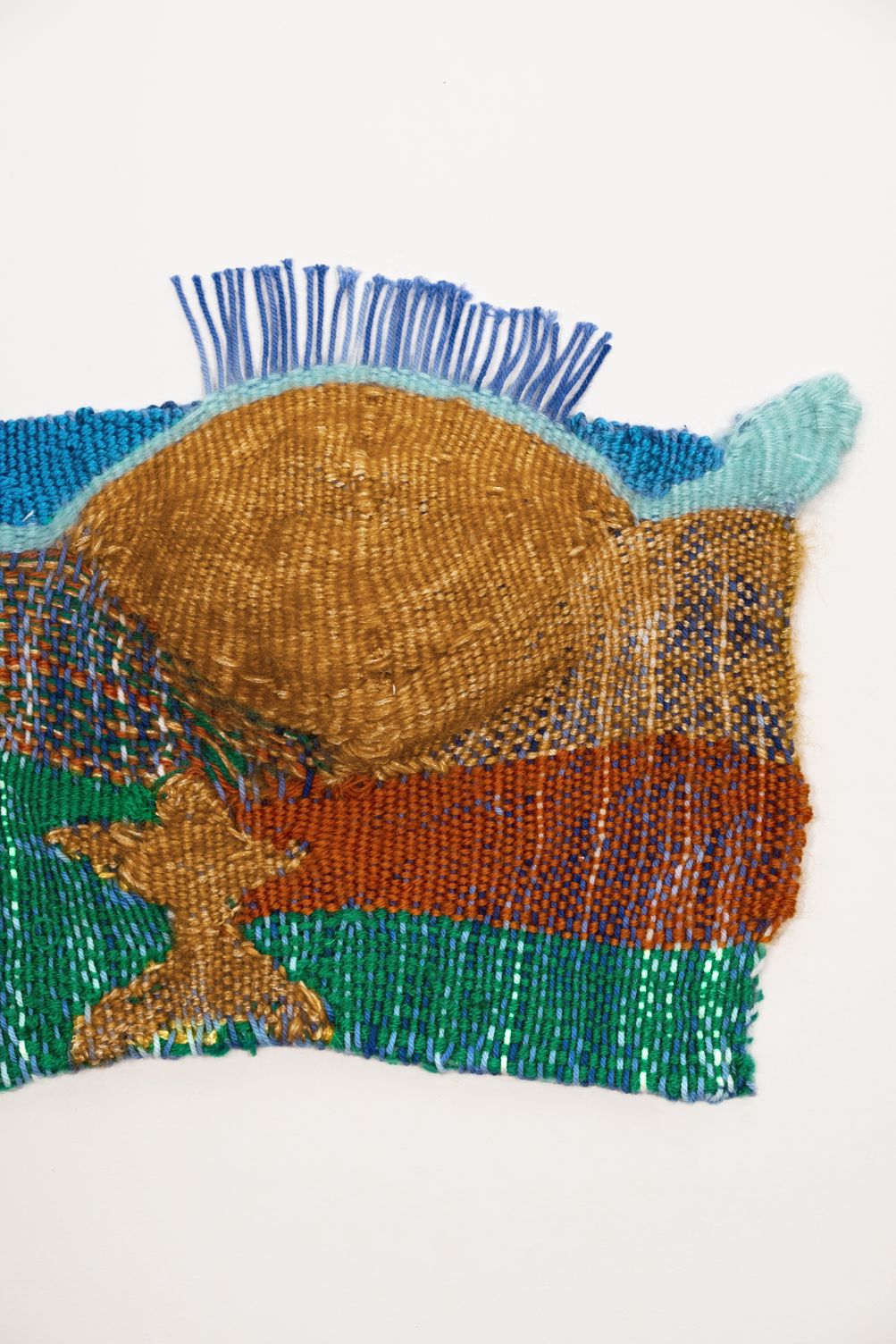 Detail of The Mountains are High: a golden woven sun behind turquoise and blue hills, above rust and green bands and a small golden figure, with a fringe of blue warp at the top
