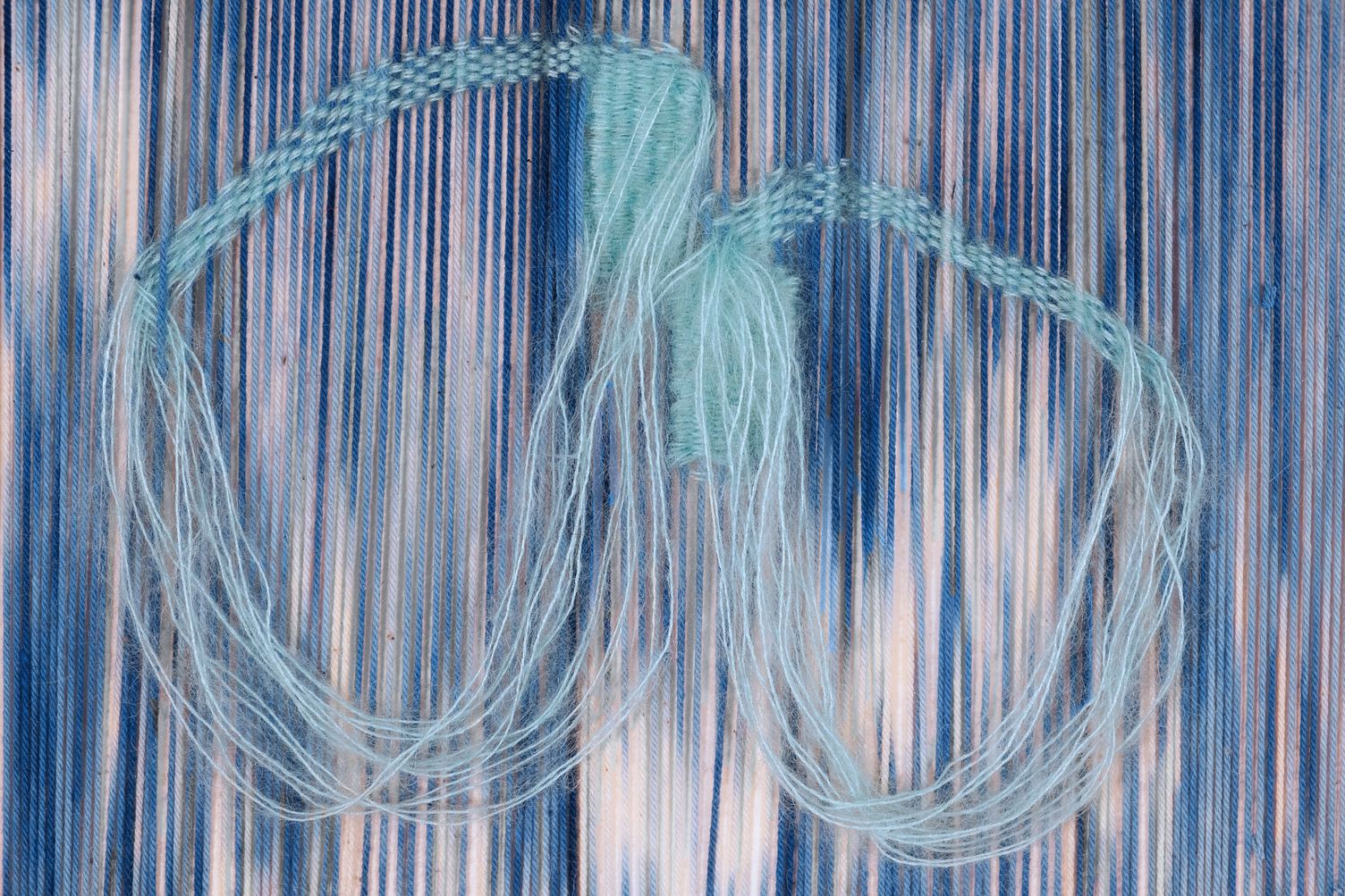 Detail of We Walk Together: two loops of pale turquoise thread, partly woven and partly hanging free, over blue and white ikat-dyed warp