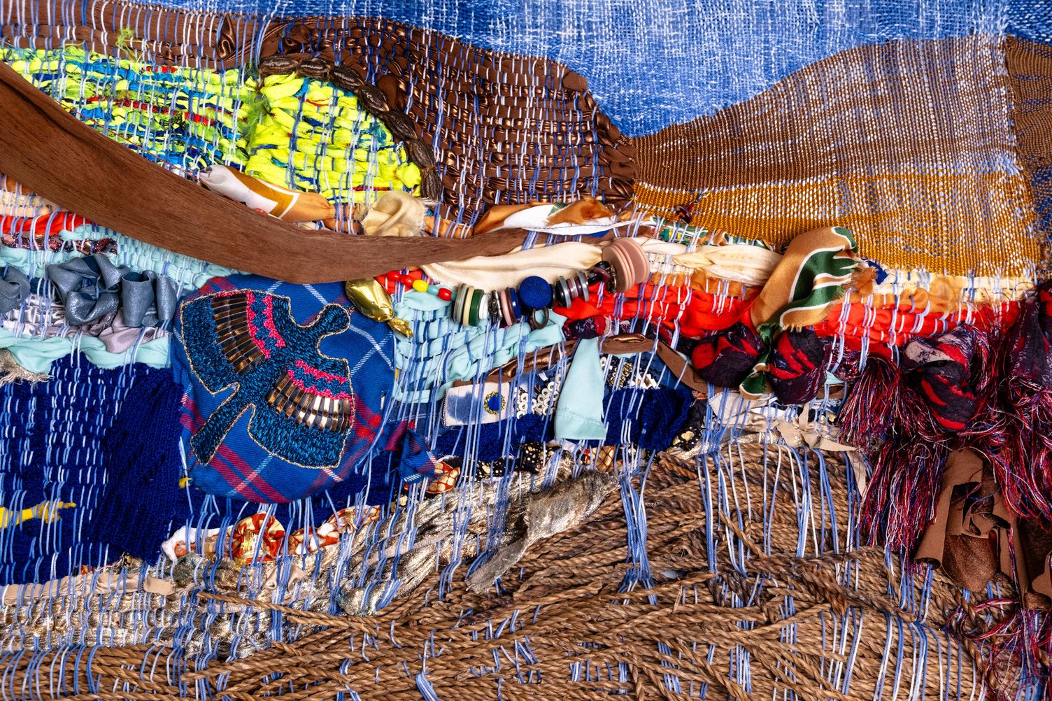 Detail of Freedom: an embroidered blue bird on tartan fabric, beneath a band of brown leather, beads and buttons, above a layer of rattan
