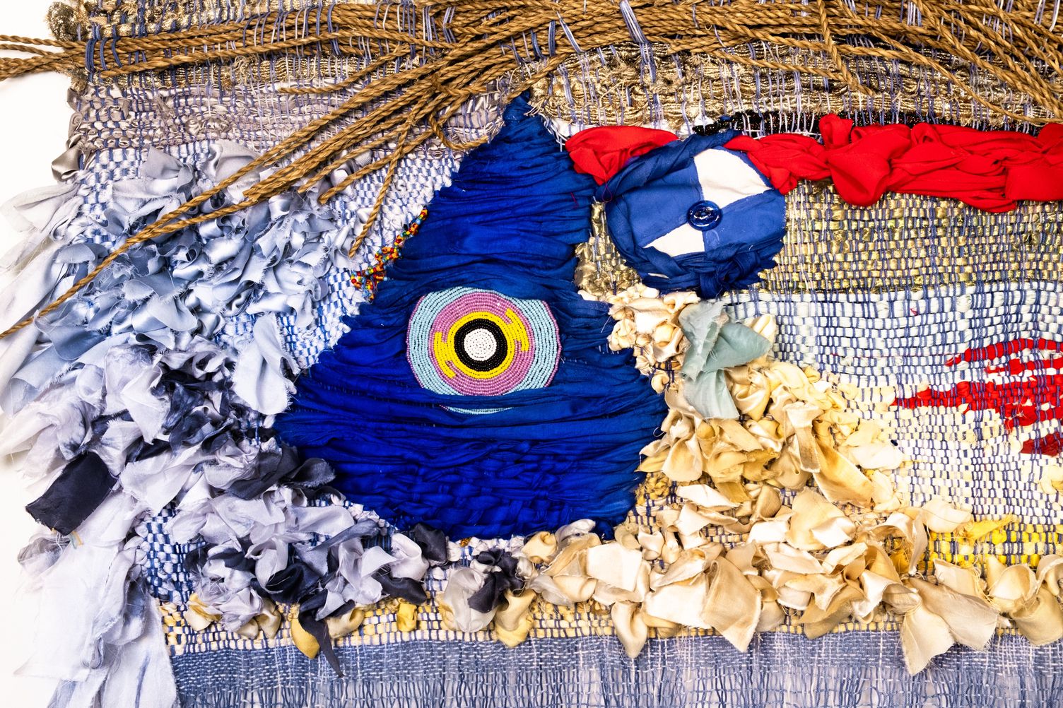 Detail of Freedom: a woven blue triangle with a beaded eye, framed by pale grey fabric strips, rattan and cream silk ribbons