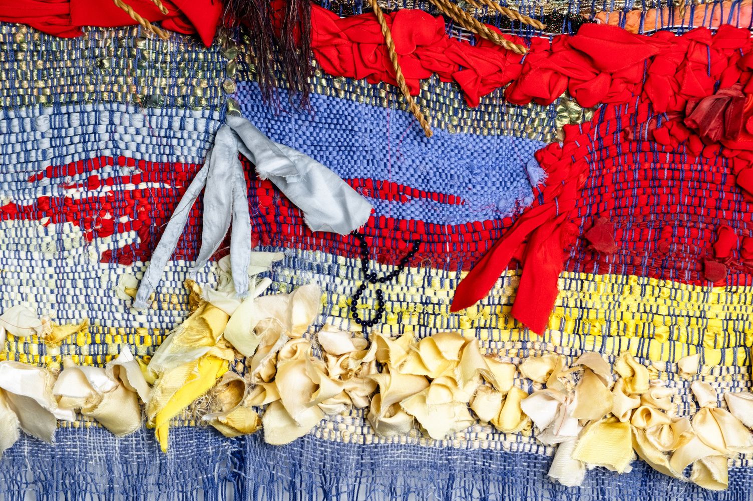 Detail of Freedom: stripes of blue, red and yellow weave with knotted red fabric and a row of cream and yellow silk ribbons