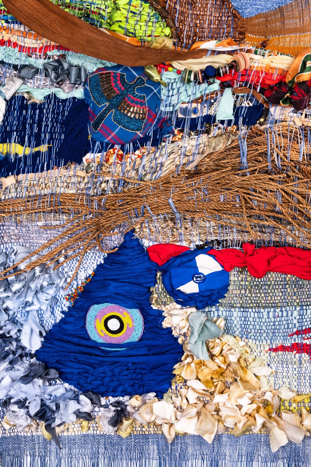 Detail of Freedom: the embroidered bird above a band of rattan, and below it the woven blue triangle with its beaded eye beside cream silk ribbons