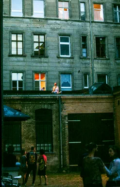 Unacknowledged Loss, page 113: colour photograph of a woman sitting cross-legged on a rooftop ledge with a rose in her mouth, singing above a courtyard of onlookers at dusk
